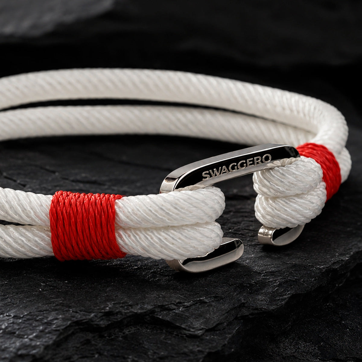 Close-up of SWAGGERO steel shackle clasp with engraved logo and screw pin, ivory rope bracelet on dark slate