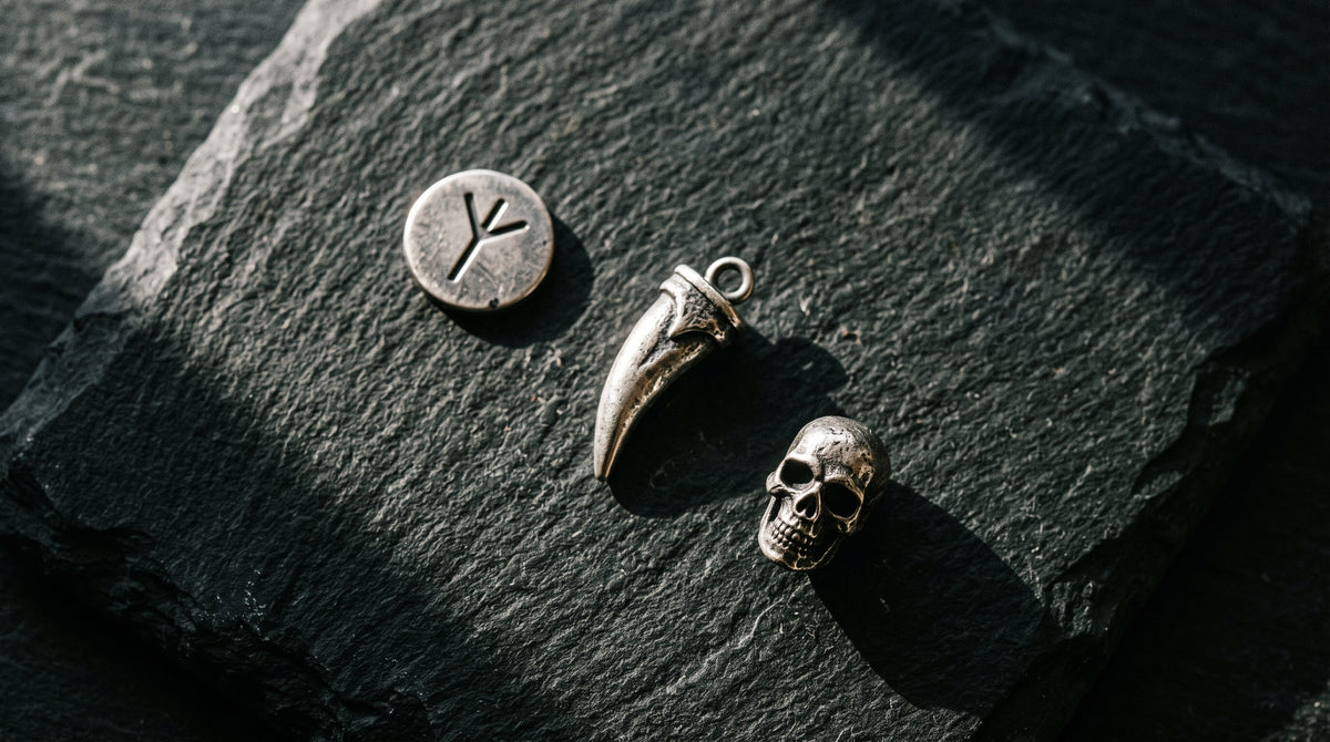 Three engraved silver bracelet charms arranged on dark slate — a Norse rune coin, a fang-shaped pendant, and a skull-shaped bead, dramatic side lighting